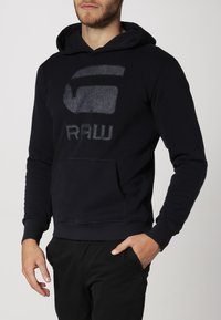 Felpa con cappuccio blu navy, dotata di una tasca frontale e un logo "RAW" testurizzato in tessuto simile al denim. Materiale di cotone morbido e liscio.