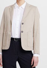 Béžový bavlněný blazer s klopovým límcem, dvěma knoflíky a dvěma předními kapsami. Nosený přes bílé tričko a pruhované kalhoty.