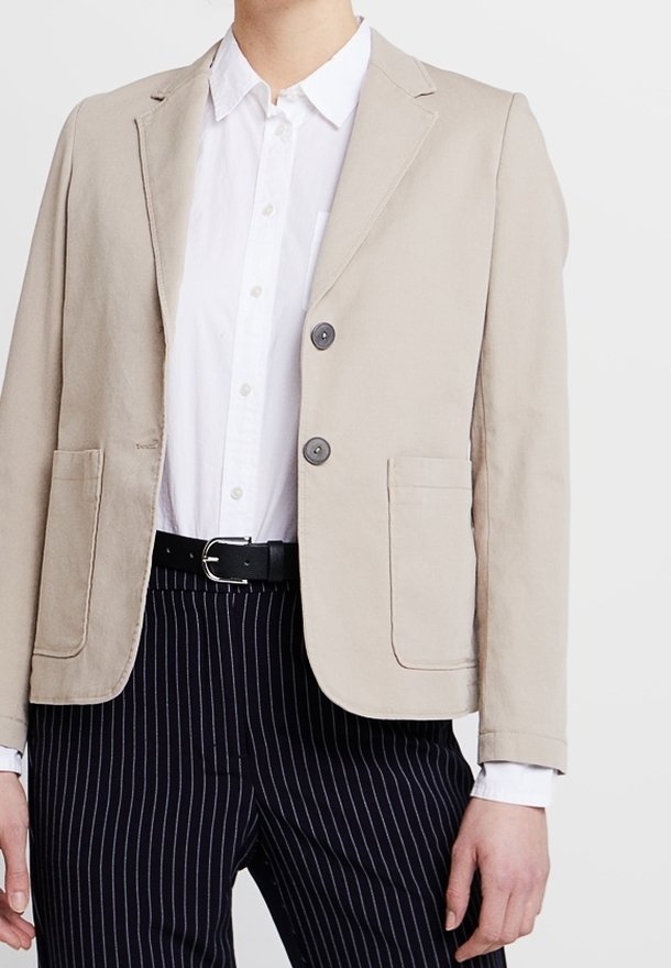 Béžový bavlněný blazer s klopovým límcem, dvěma knoflíky a dvěma předními kapsami. Nosený přes bílé tričko a pruhované kalhoty.