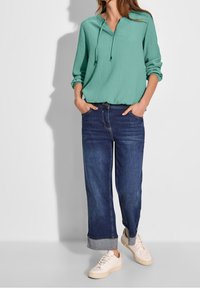 Blouse turquoise à coupe décontractée, manches longues et capuche ; associée à un jean bleu foncé à jambes larges et des baskets beige clair.