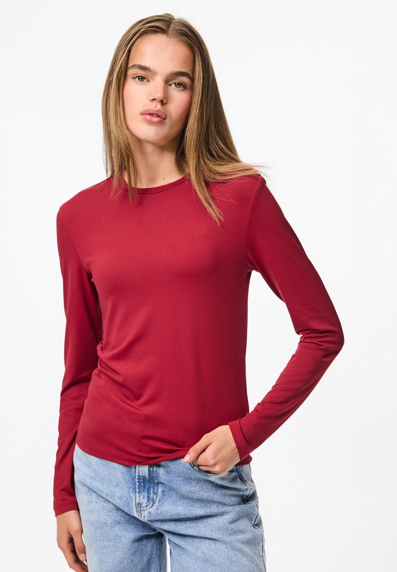 Jonge vrouw met steil blond haar draagt een aansluitend rood shirt met lange mouwen en lichtblauwe jeans, staande met één hand in haar zak.
