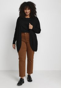 Schwarze gestrickte Strickjacke mit offenem Vorderteil, getragen über einem schwarzen Oberteil und braunen hochgeschnittenen Hosen. Schwarze Slipper runden das Outfit ab.