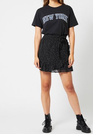 T-shirt noir avec le texte "NEW YORK" en bleu, associé à une jupe cache-cœur noire à pois blancs avec un ourlet froncé et des bottes noires.