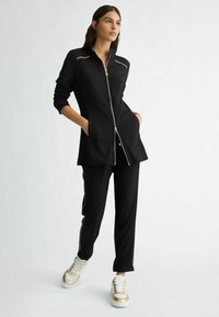 Črna športna obleka z zip-up jakno s srebrnimi detajli in usklajenimi hlačami s stranskimi črtami. Model nosi zlate superge.