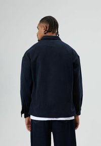 Man met gevlochten haar, gekleed in een donkerblauwe jas en donkere spijkerbroek, staand met de rug naar voren tegen een effen lichte achtergrond.