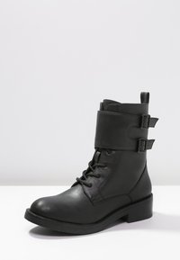 Bota de cuero negro hasta el tobillo con puntera redonda, cordones en la parte delantera y dos correas ajustables con hebillas alrededor de la parte superior. Suela de goma plana.