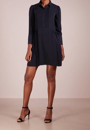 Kvinde iført en marineblå langærmet kjole med plisserede detaljer foran og sorte hæle med ankelrem, stående på en neutral gulv.