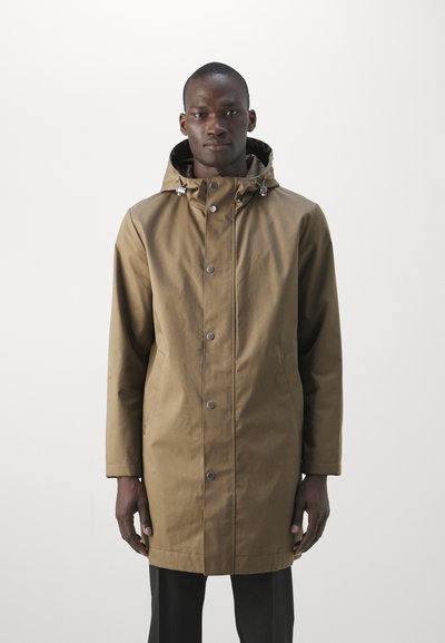 Uomo che indossa un cappotto marrone a maniche lunghe con cappuccio e bottoni a scatto sopra pantaloni neri, in piedi su uno sfondo bianco semplice.