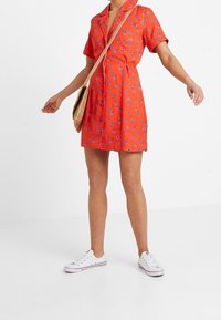 Röd klänning med korta ärmar och blå blommönster, knappstängning fram, justerbart midjeband och avslappnad passform. Bärs tillsammans med vita sneakers och en vävd väska.