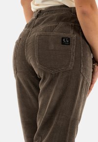 Pantaloni in corduroy marroni con texture a coste, dotati di tasche posteriori e una patch nera con logo. La vestibilità su misura accentua la forma.