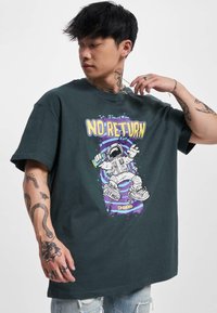 Camiseta de algodón verde oscuro con un estampado gráfico de un astronauta y el texto "NO HAY RETORNO" junto a salpicaduras coloridas en la parte frontal.