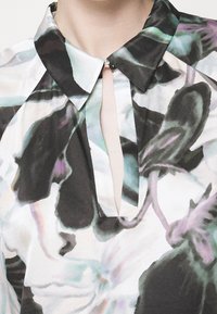 Blouse à col avec un motif floral abstrait en noir, blanc et pastel, dotée d'une ouverture cruciforme devant, fermée par un petit bouton.