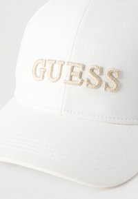 Λευκό καπέλο μπέιζμπολ με ανάγλυφο, μπεζ κεντημένο λογότυπο "GUESS". Το υλικό έχει λείο ύφασμα με ορατές ραφές.