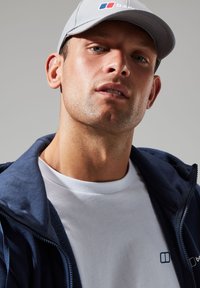 Navy zip-up jacket over a white t-shirt, gray cap with logo. Close-up on face with short hair and light stubble, neutral background.