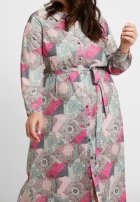 Robe longue à boutons, fabriquée dans un tissu présentant un design en patchwork dans des tons de rose, gris et vert, avec des motifs complexes et une ceinture à la taille.