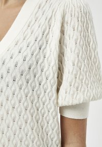 Strickpullover in Creme mit einem wellenartigen Muster, V-Ausschnitt und kurzen Ärmeln mit gerippten Bündchen. Leichtes, strukturiertes Material.