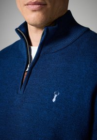 Dunkelblaues Zip-Up-Sweatshirt mit hohem Kragen, texturiertem Strickstoff und kleinem, hell gefärbtem Reh-Logo auf der Brust. Sichtbarer silberner Reißverschluss.