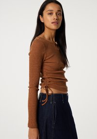 Brown ribbed long-sleeve top with gathered sides, featuring a keyhole detail at the neckline and ties, paired with a dark denim skirt.