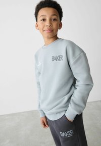 Lys grå sweatshirt med ribbede manchetter og kant, med "BAKER" logo i mørkegrå. Sammen med mørkegrå bukser, der viser det samme logo.
