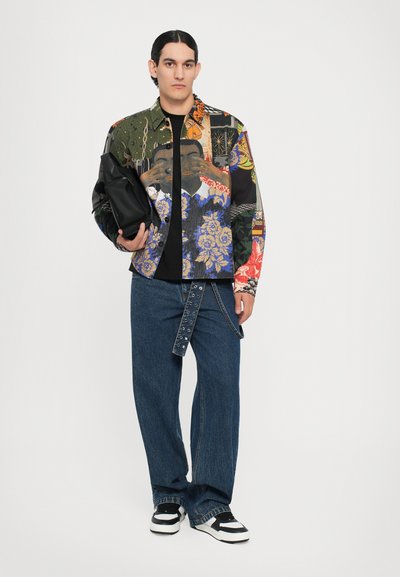 Bunt gemustertes Hemd mit verschiedenen Motiven, schwarzes T-Shirt, weite blaue Jeanshose und schwarz-weiße Sneakers. Hält eine schwarze Tasche.