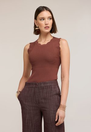 Vrouw met een mouwloze roestkleurige top met gegolfde randen en een hoge taille bruine broek met textuur, geaccentueerd met gouden sieraden.