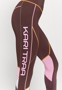 Leggings sportivi marroni con accenti rosa e gialli, pannelli in rete e il testo "KARI TRAA" in rosa lungo la coscia esterna.