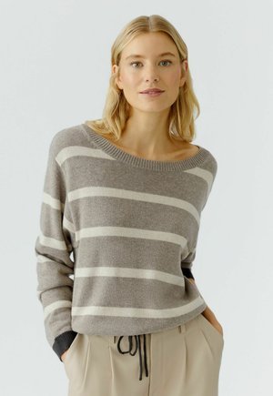 Blonde vrouw in een grijs-wit gestreepte trui met zwarte manchetten en beige broek, staand met de handen in de pockets tegen een effen achtergrond.