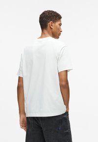 Vīrietis ar cirtainiem matiem, tērpies vienkāršā baltā īsām piedurknēm t-kreklā un melnās džinsās, stāv ar muguru pret kameru.