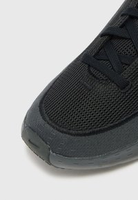 Sapatilha desportiva preta com um upper em malha texturizada e sola de borracha lisa. Os atacadores são pretos, com uma marcação discreta visível na ponta.