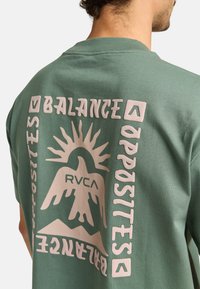 Green cotton t-shirt featuring large pink text and graphic of a mountain with a sun, displaying the words "Balance" and "Opposites."
