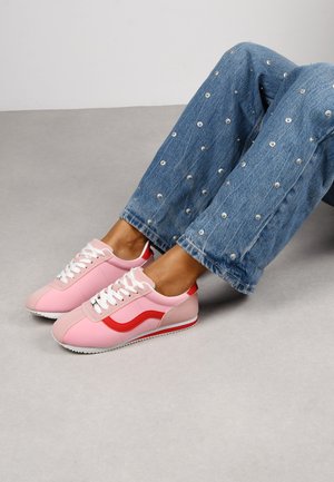 Zapatillas rosas con acentos rojos, con cordones blancos y una suela texturizada. Llevadas con unos jeans de mezclilla azules adornados.