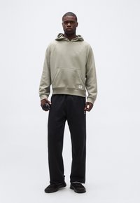 Άνδρας που στέκεται μπροστά από απλό φόντο, φοράει ανοιχτό μπεζ φούτερ με κουκούλα, μαύρο φούτερ παντελόνι, μαύρα αθλητικά παπούτσια, κρατώντας ένα μαύρο παγούρι νερού στο αριστερό χέρι.