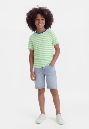 Uśmiechnięte dziecko z kręconymi włosami, ubrane w zielono-białą koszulkę w paski, jasne jeansowe szorty i białe trampki, stoi z jedną ręką w kieszeni.