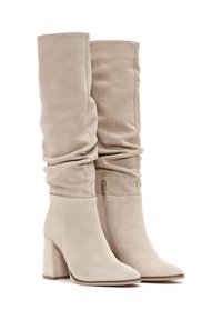 Beżowe zamszowe kozaki na wysokim obcasie o luźnym fasonie, z szpiczastym noskiem i krótkim zapięciem bocznym. Charakteryzują się blokowym obcasem i miękką teksturą.