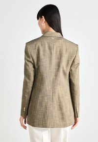 Beige rakenne blazer, jossa on muokattu istuvuus, yksittäinen halkio takana ja kultaisella värillä varustetut napit hihoissa. Siistit linjat ja klassinen muotoilu.