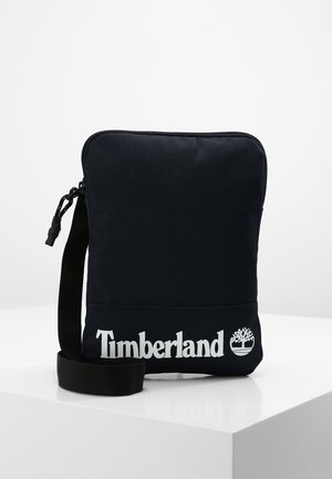 Zwarte rechthoekige crossbodytas met ritssluiting en verstelbare band, met wit Timberland-logo en boomembleem aan de voorkant onderaan.