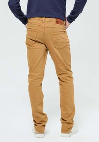 Jeans beige slim fit avec une texture lisse, cinq poches, et une étiquette en cuir marron sur la ceinture arrière. Baskets blanches visibles en dessous.