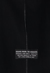 Zwarte stof met een verticale naad met metalen stiksel. Label met de tekst "ESCAPE FROM / THE UNKNOWN," met aanvullende tekst eronder.