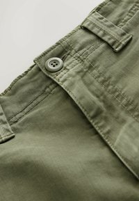 Gros plan sur la ceinture d'un pantalon vert olive montrant le bouton, les passants de ceinture et les coutures détaillées sur le tissu.