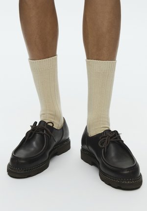 Nohy v krémových žebrovaných crew ponožkách a čiernych kožených šnurovacích topánkach s hrubými podrážkami na bielom pozadí.