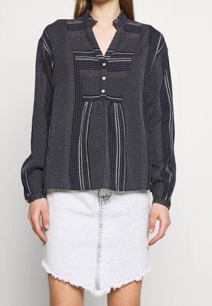 Blouse noire à motifs avec un col mandarin, fermeture par boutons et manches longues. Associée à une mini jupe en jean blanche avec un ourlet effiloché.