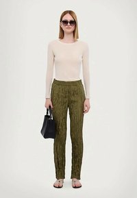 Nainen, joka on pukeutunut vaaleanbeigeen pitkähihaiseen puseroon, oliivinvihreisiin teksturoituihin housuihin, mustaan käsilaukkuun, aurinkolaseihin ja sandaaleihin, yksinkertaista valkoista taustaa vasten.