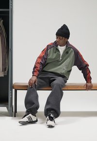 Sportinė striukė alyvuogių žalios spalvos su bordo ir oranžiniais akcentais, derinama su pilkomis sportinėmis kelnėmis. Juoda megzta kepurė ir dryžuoti juodos ir baltos spalvų sportbačiai.