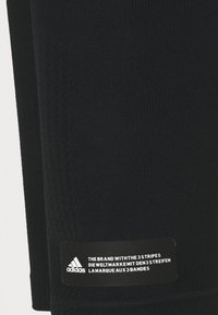 Černé žebrované legíny s texturovaným povrchem, které mají obdélníkovou nášivku loga Adidas s vícejazyčným brandingem.