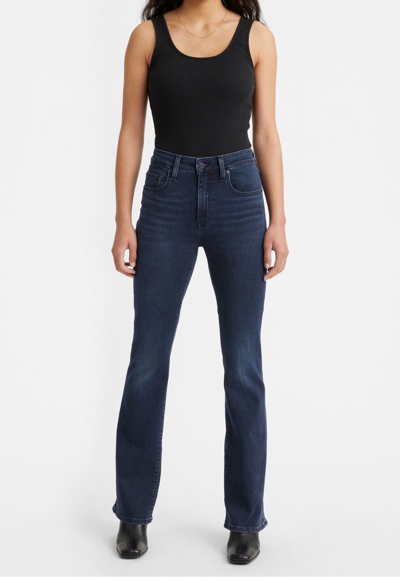 Top senza maniche nero a costine abbinato a jeans a zampa d'elefante blu scuro. I jeans hanno cinque tasche e una chiusura lampo classica. Le calzature sono stivali neri con tacco.