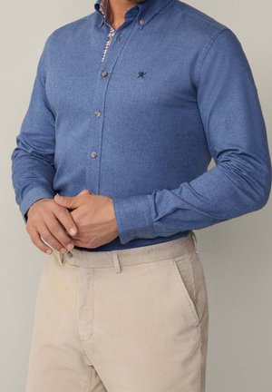 Homme aux mains croisées portant une chemise boutonnée à manches longues bleue et un pantalon beige, sur un fond neutre.