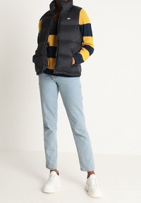 Czarna kamizelka puchowa noszona na żółto-granatowym swetrze w paski, z jasnoniebieskimi dżinsami i białymi trampkami. Zawiera zamek błyskawiczny i kieszenie boczne.