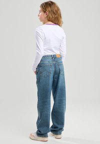 Jeans in denim blu con una vestibilità ampia, dotati di un alto giro vita, grandi tasche posteriori e un'etichetta in pelle marrone. Abbinate a una camicia bianca a maniche lunghe.