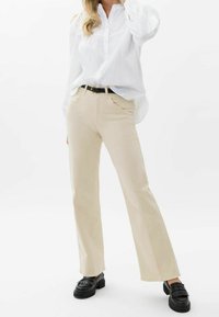 Pantalones de campana beige con bolsillos frontales y una textura suave, combinados con una camisa blanca de botón y mocasines negros con detalles de flecos.