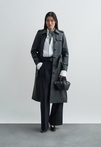 Svart trenchcoat i läder med knäppning framtill, kombinerad med en vit skjorta, grå slips och svarta byxor med vida ben. Accessoarer inkluderar en liten svart handväska.